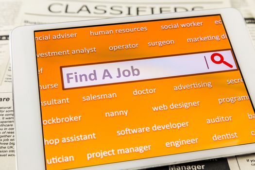 Orange tablet screen fill with difference career words. Internet website for online job search having wording "find a job" and searching symbol in search engine box. Blur classifieds ads on background. Top view image.