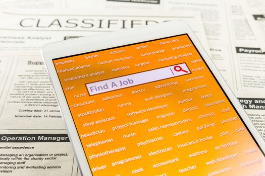 Orange tablet screen fill with difference career words. Internet website for online job search having wording "find a job" and searching symbol in search engine box. Blur classifieds ads on background. Bird eye view image.