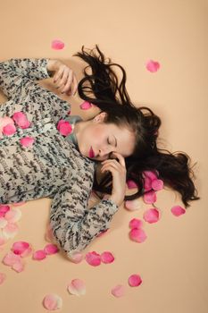 atractive brunette girl lying on beige background, dreaming and sleeping with rose petals around. Beaty concept.