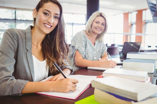 Smiling matures females students writing notes at desk in library