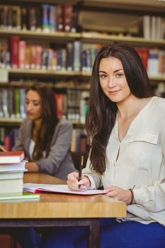 Happy student sitting at desk writing smiling at camera in library