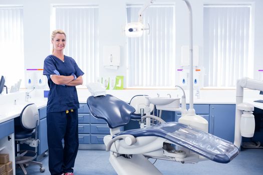 Dentist in blue scrubs standing with arms crossed beside chair at the dental clinic