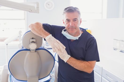 Smiling dentist leaning against dentists chair in dental clinic