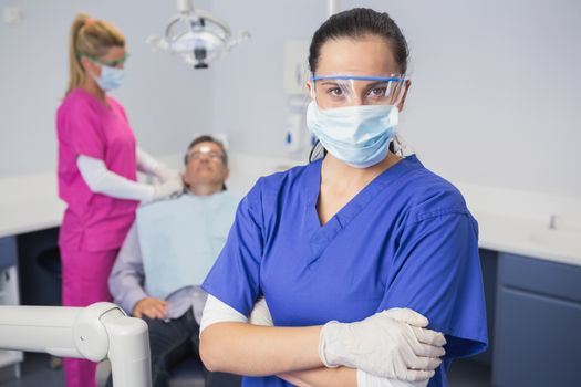 Dentist wearing surgical mask and safety glasses arms crossed behind her patient in dental clinic
