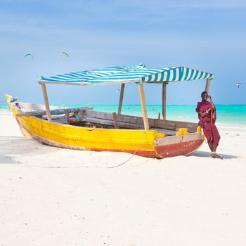 Maasai warrior lounging aroundon traditional colorful wooden boat on picture perfect tropical sandy beach on Zanzibar, Tanzania, East Africa. Kiteboarding spot on Paje beach.