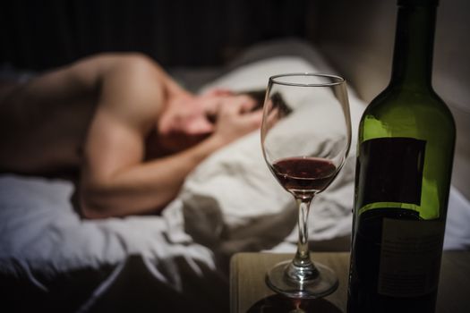 Hangover Man with Headaches in a Bed at Night and Wine bottle in Focus