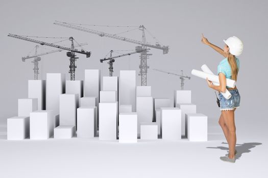 Beautiful girl in working clothes holding paper scrolls and pointing finger. Rear view. Minimalistic city of white cubes with wire-frame tower cranes on gray background
