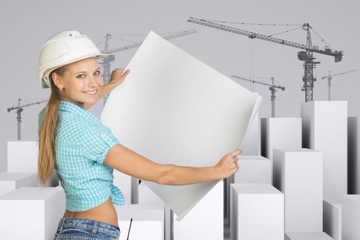 Beautiful girl in white helmet showing empty paper sheet, looking at camera, smiling. Minimalistic city of white cubes with wire-frame tower cranes on gray background