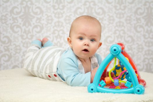 Blue eyes baby girl lying on belly on soft surface , playing with colourful toy and looking at the camera