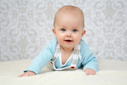 Cute  baby girl with blue eyes laying on belly on soft surface, wearing blue vest and striped dungarees. Looking at the camera