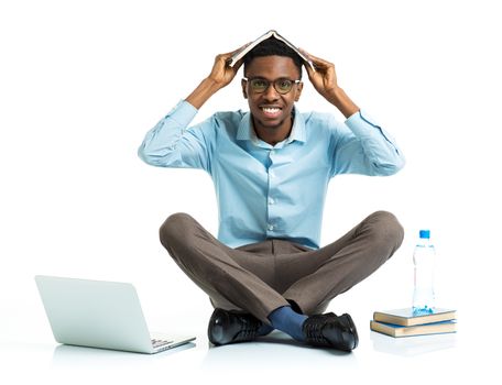 Happy african american college student sitting with laptop and book on his head on white background
