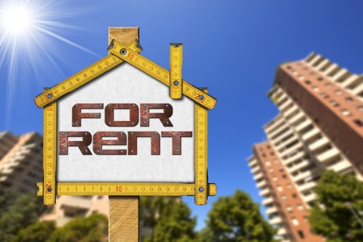 Yellow wooden meter ruler in the shape of house with text for rent. For rent real estate sign with tall and blurred buildings in the background