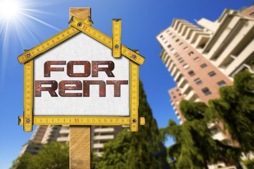 Yellow wooden meter ruler in the shape of house with text for rent. For rent real estate sign with tall and blurred buildings in the background