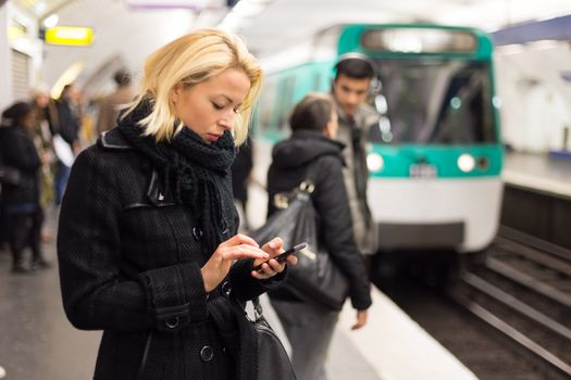 Young woman in winter coat with a cell phone in her hand waiting on the platform of a railway station for train to arrive. Public transport.  