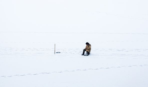 Fisherman sitting near the ice-hole on the frozen water surface covered with snow. 