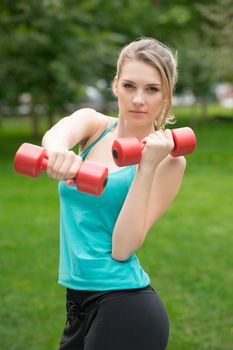 Young beautiful sports girl with dumbbells in the park on a background of green grass