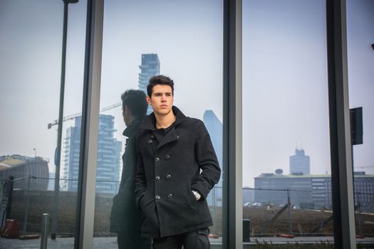 Stylish trendy young man standing outdoor against office window, looking confindent away