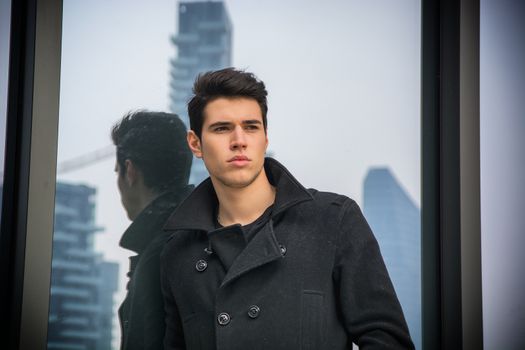 Stylish trendy young man standing outdoor against office window, looking confindent away