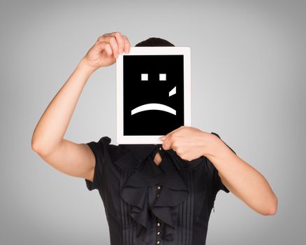 Girl in dress covered her face with tablet. On screen sad smiley. Gray background