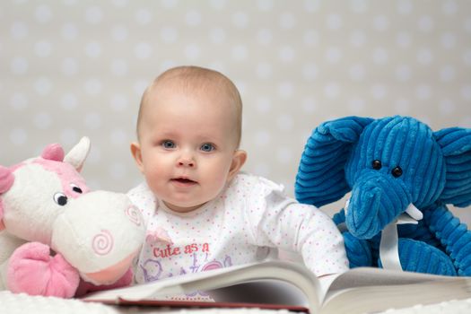 Baby girl reading a book with little toy friends and lookin at the camera