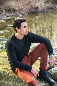 Attractive young man sitting outdoor in nature, at river or water stream, looking away to a side