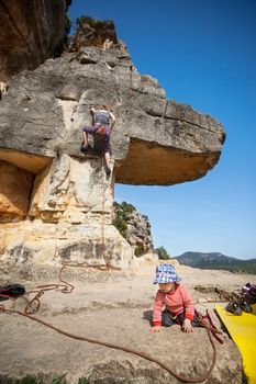 Toddler boy playing at foot of mountain while his mother climbing at the background. The boy is tied in to the rope.