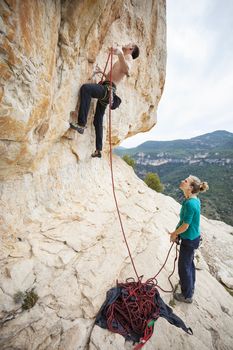 Rock climber going to clip rope at beginning of route, girl belayer watching him