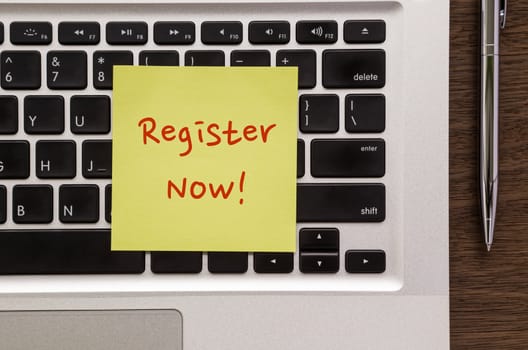 Top view of yellow notepad with words " Register Now " putting on laptop keyboard and stainless steel pen on wood table