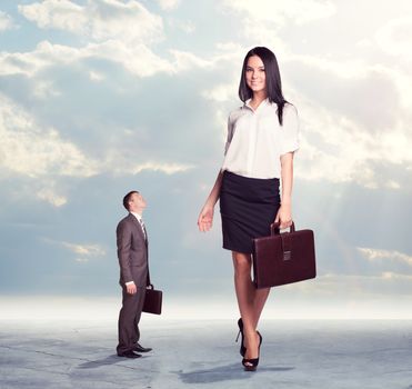 Small businessman looking up at on high walking businesswoman. Clouds and cement surface as background. Business concept