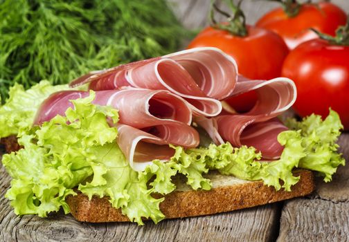 Sliced prosciutto with lettuce, bread, tomatoes and herb on wooden background