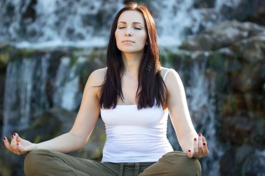 Young woman in ltus pose sitting near watefall