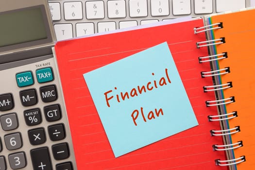 Close up blue notepad with words " Financial Plan " putting on on colorful paper book with keyboard and calculator on background
