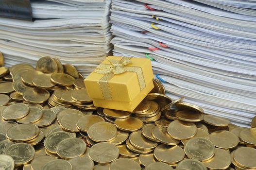 Gift box in heap of gold coins with pile of paperwork.                               