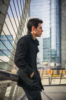 Profile of Stylish Young Handsome Man in Black Coat Standing in City Center Street with Skyscraper Behind Him, Looking to the Right of the Frame.