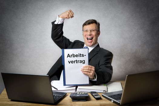 Happy businessman at the office desk is holding a employment contract with german title "Arbeitsvertrag"