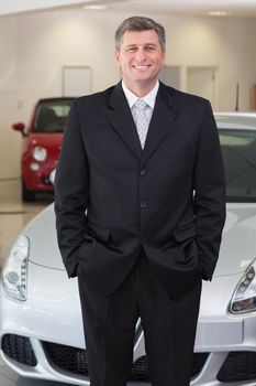 Smiling businessman standing with hands in pockets at new car showroom