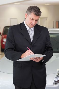 Serious businessman writing on clipboard at new car showroom