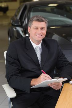 Businessman sitting at his desk while writing on clipboard at new car showroom