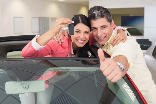 Smiling couple giving thumbs up and holding car key at new car showroom