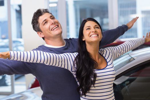 Happy couple standing with arms outstretched at new car showroom