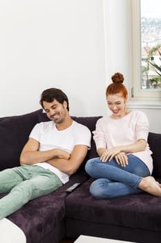 Happy Young Sweet Couple in Casual Clothing Smiling at Each Other While Sitting at the Couch in the Living Room