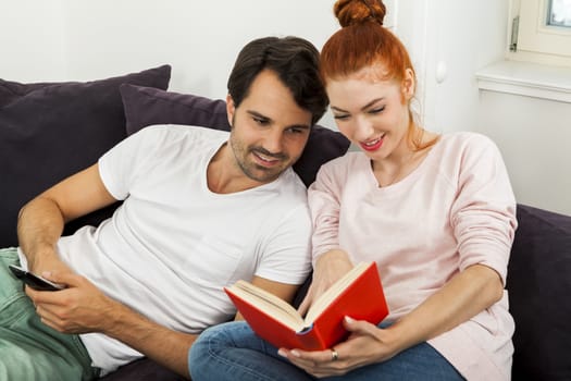 Young Couple Resting on the Sofa at the Living Room While the Man is Watching TV and Woman is Reading a Book.
