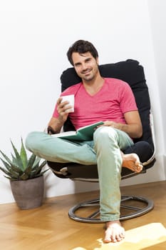 Young Man in Casual Clothing Sitting on Black Chair While Reading a Book and Holding a Glass of Drink.