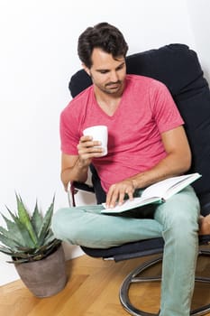 Young Man in Casual Clothing Sitting on Black Chair While Reading a Book and Holding a Glass of Drink.