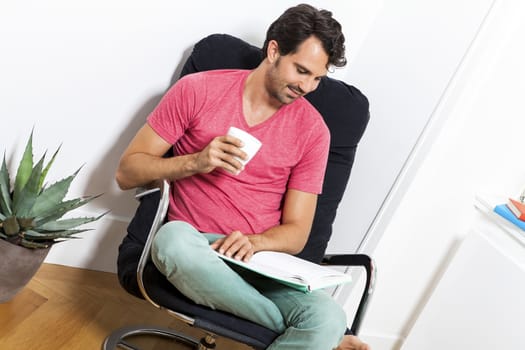 Young Man in Casual Clothing Sitting on Black Chair While Reading a Book and Holding a Glass of Drink.