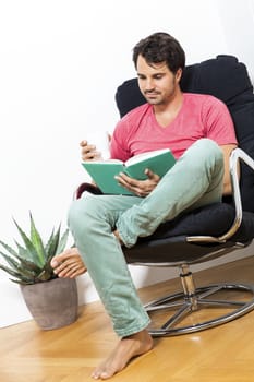 Young Man in Casual Clothing Sitting on Black Chair While Reading a Book and Holding a Glass of Drink.