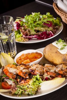 Grilled prawns with a green leafy lettuce and endive salad and a jacket potato topped with sour cream served on a white plate, close up high angle view on white