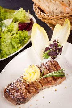 Tasty grilled beef steak topped with a twirled knob of butter and a sprig of fresh rosemary and served on a white plate, close up view