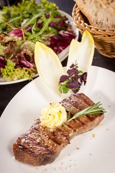 Tasty grilled beef steak topped with a twirled knob of butter and a sprig of fresh rosemary and served on a white plate, close up view