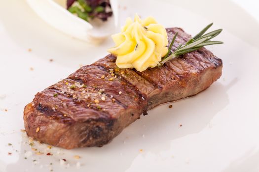 Tasty grilled beef steak topped with a twirled knob of butter and a sprig of fresh rosemary and served on a white plate, close up view